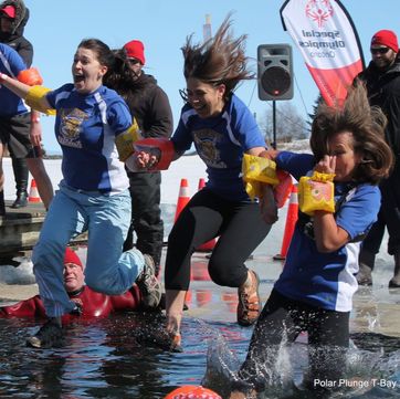 Three women in school tshirts and water wings jump into a hole in the frozen lake