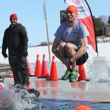 A man wearing a bright orange swim cap does a cannon ball dive into a hole in the frozen lake. 