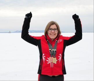 A female athlete stands on the frozen lake wearing her special olympics uniform, two medals around her neck with her hands in the air. 